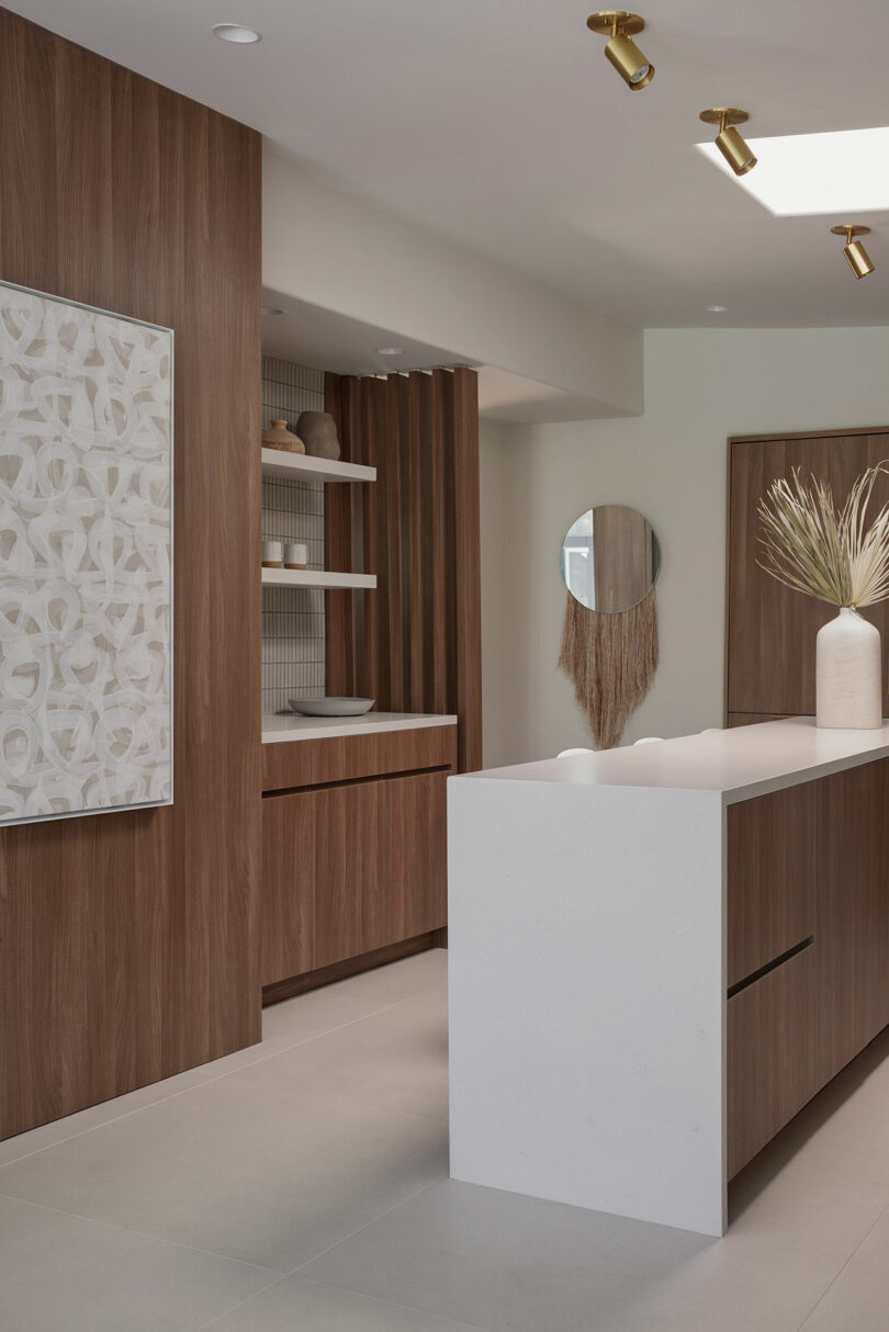 Modern kitchen with wood cabinets, a white island, and minimalist decor. A round mirror hangs on the wall, and a vase with pampas grass sits on the island.