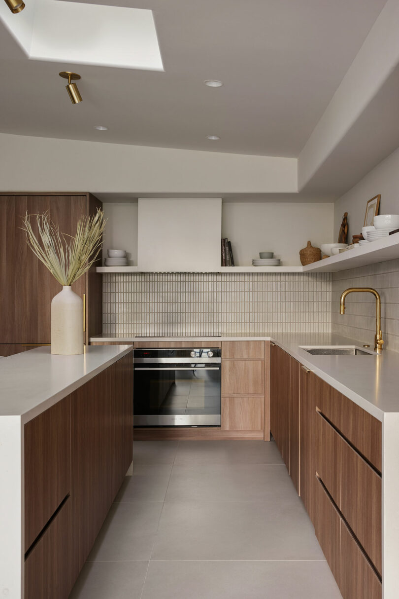 Modern kitchen with wood cabinets, white countertops, and a gold faucet. Oven is built into cabinetry. White tile backsplash and decorative vase on the countertop.