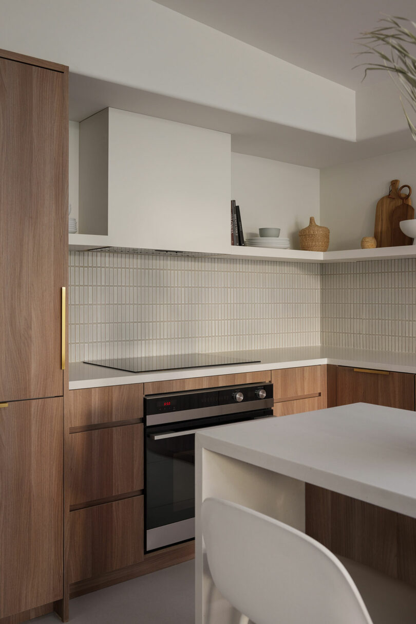 Modern kitchen with wooden cabinets, white countertops, built-in oven, and tiled backsplash. Open shelving holds decor items like bowls and cutting boards. Minimalist and clean design.