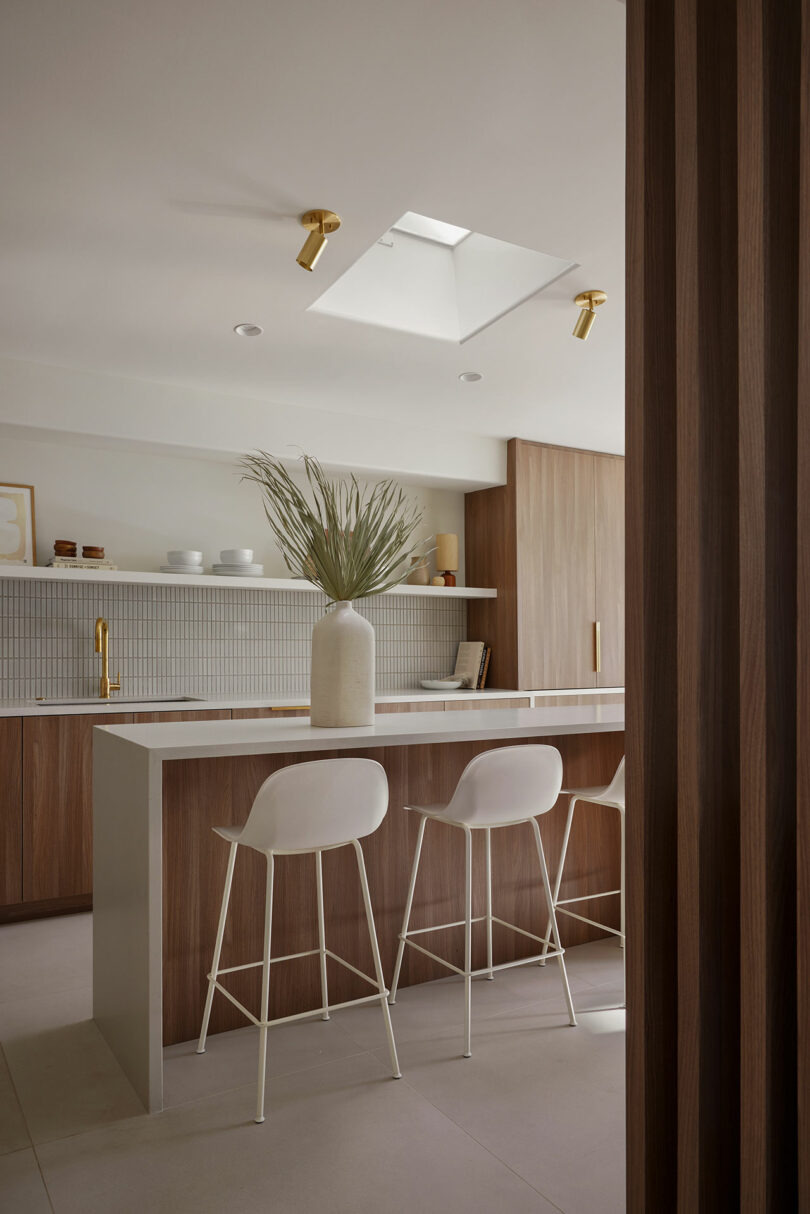 Modern kitchen with wooden cabinets, a central island with two white stools, and a large vase with greenery. A skylight and minimal decor create a bright, open space.