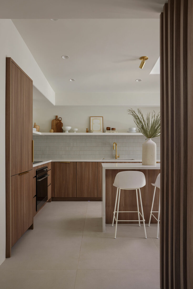 Modern kitchen with wood cabinetry, a white countertop, bar stools, and minimal decor. Neutral color palette with a potted plant on the counter and open shelving displaying ceramics.