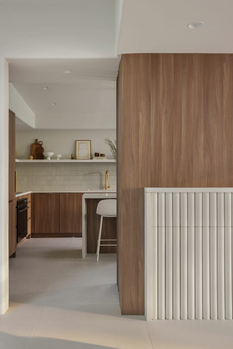 Modern kitchen with wood cabinetry, a white countertop, bar stool, and beige tiled backsplash. Neutral tones create a minimalist look.