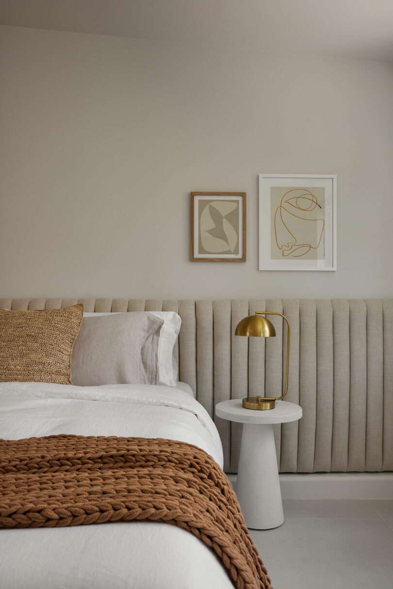 A modern bedroom with a beige upholstered headboard, white bedding, a brown knitted throw, a gold lamp on a round white side table, and two abstract artworks on the wall.