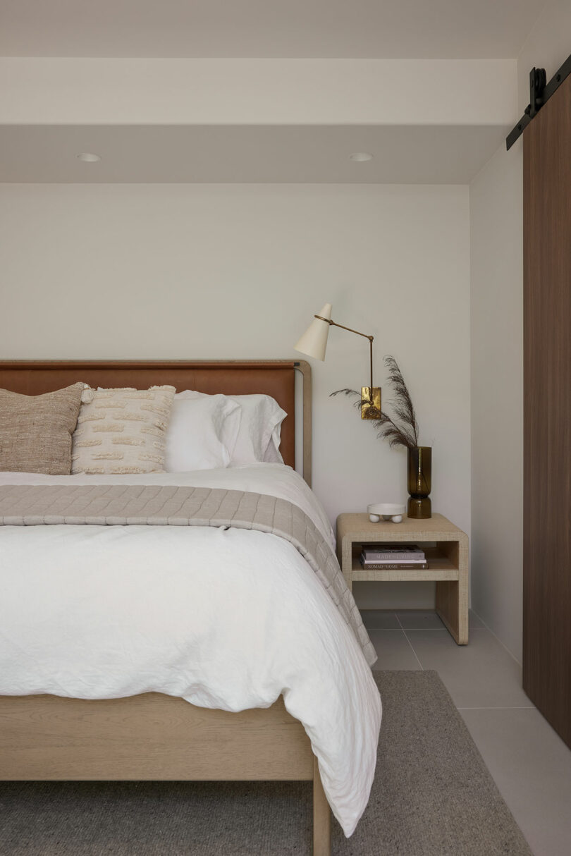 Minimalist bedroom with a white bed, beige pillows, a wooden nightstand with a vase and lamp, and a sliding wooden door on the right.