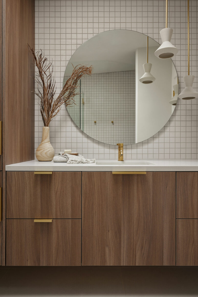 Modern bathroom with a round mirror, wooden cabinets, gold handles, and pendant lights. A vase with dried branches is on the countertop, set against a tiled wall backdrop.