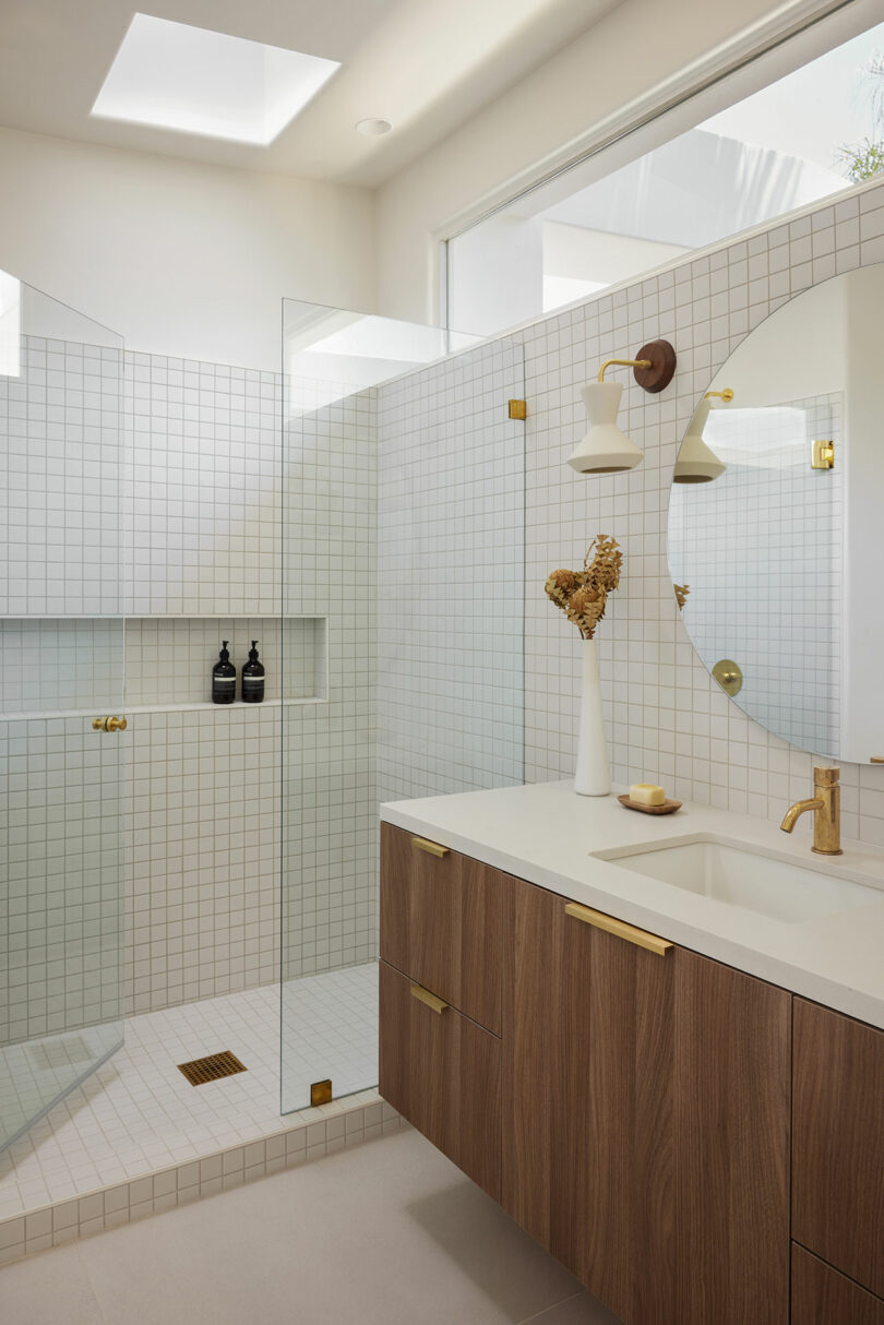 Modern bathroom with a wooden vanity and round mirror. Glass shower enclosure with white square tiles. Two soap dispensers and a decorative vase with dried flowers on the counter.