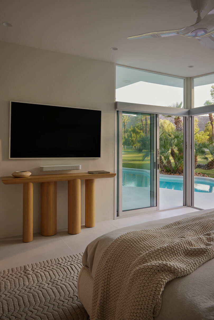 A modern bedroom with a large bed, wall-mounted TV, and a view of a pool and garden through sliding glass doors.