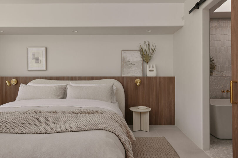 Modern bedroom with a neutral color palette, featuring a bed, wooden headboard, wall art, and a side table. A bathroom with a bathtub is partially visible through a sliding door.