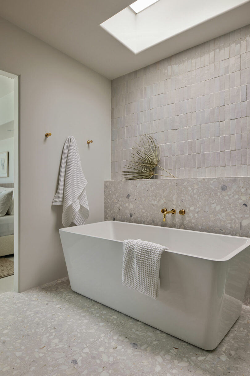 Minimalist bathroom with large white bathtub, gold fixtures, light stone tile wall, and terrazzo flooring. A towel hangs on the tub and another on a wall hook. Ceiling skylight present.