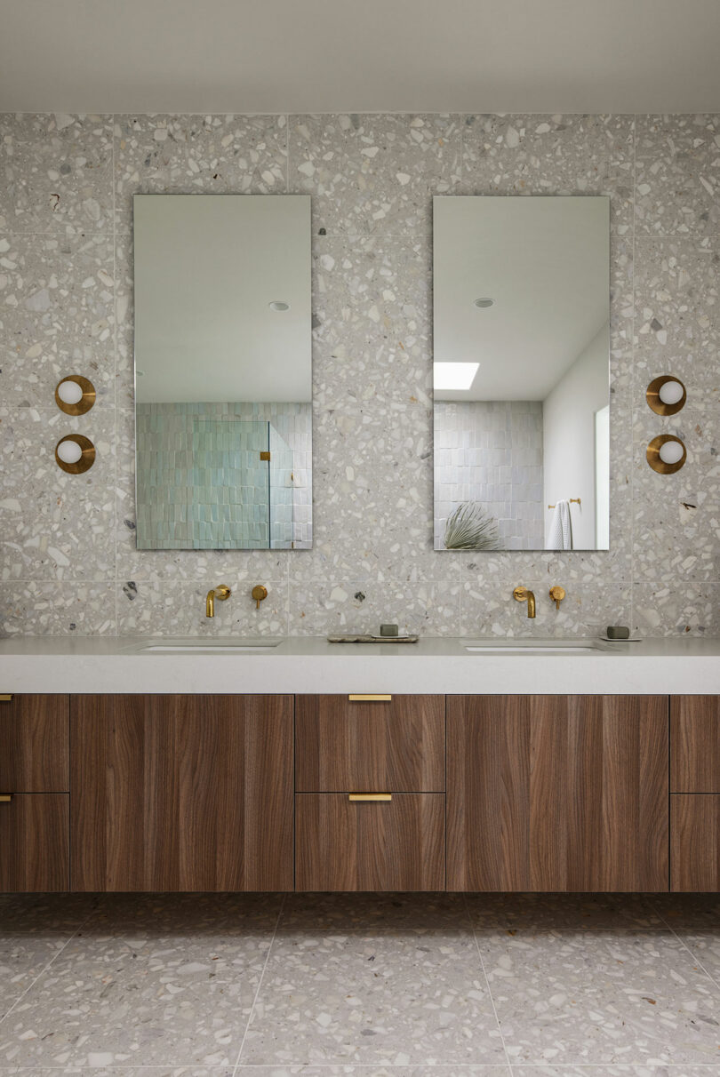 Modern bathroom with a large vanity, dual mirrors, terrazzo walls, and brass fixtures. Light wooden cabinetry and a tiled shower area are visible.