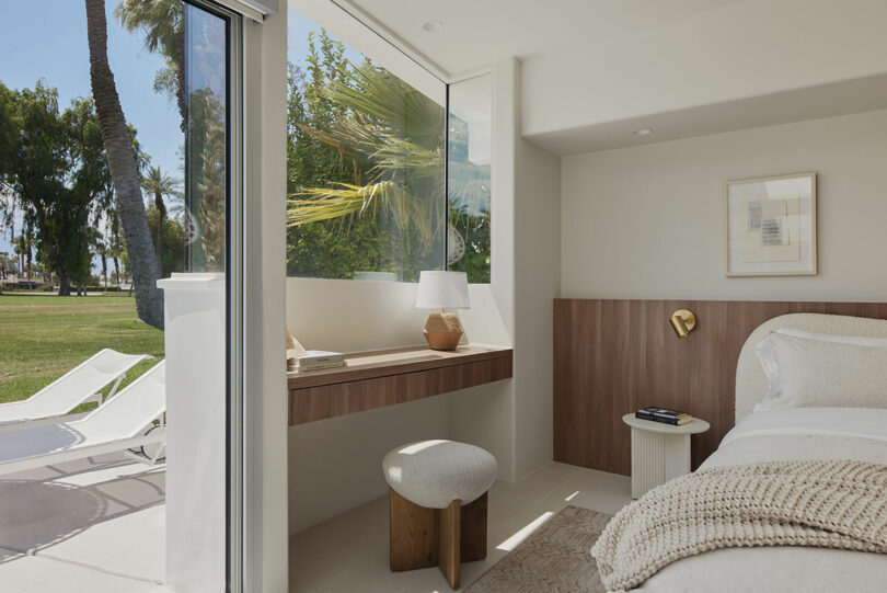 Modern bedroom with large windows, a desk with a lamp, a stool, and a bed. Views of palm trees and white lounge chairs outside.