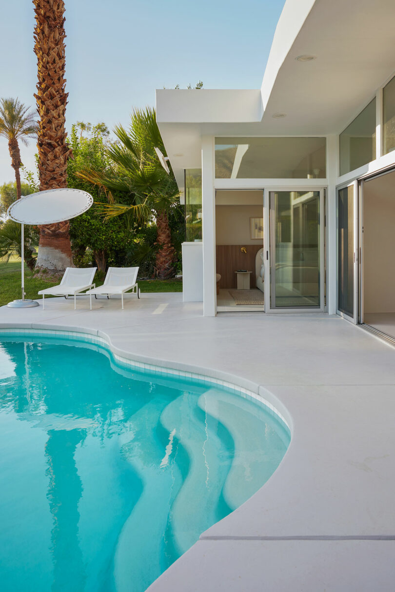 Modern house with a glass door opening to a patio featuring a pool, two lounge chairs, and surrounded by palm trees.