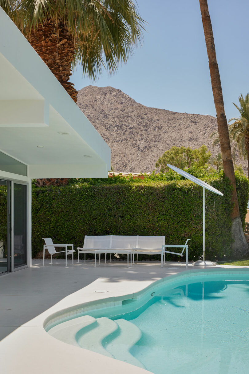 Modern backyard with a swimming pool, white patio furniture, a tall palm tree, and a mountain in the background under a clear blue sky.