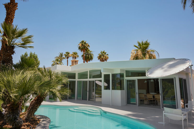 A modern house with large windows, surrounded by palm trees, features a swimming pool in the foreground under a clear blue sky.