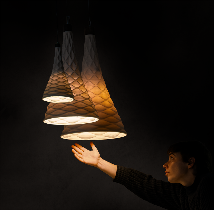 A person reaches towards three illuminated, textured pendant lamps in a dimly lit room