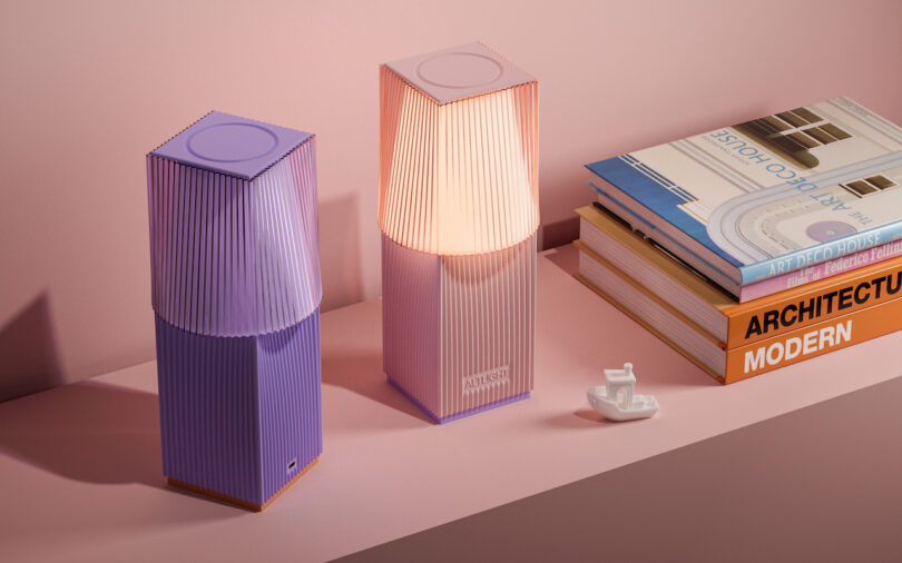 Two geometric table lamps, one lit and one not, on a pink surface next to a stack of modern architecture books and a small ceramic object