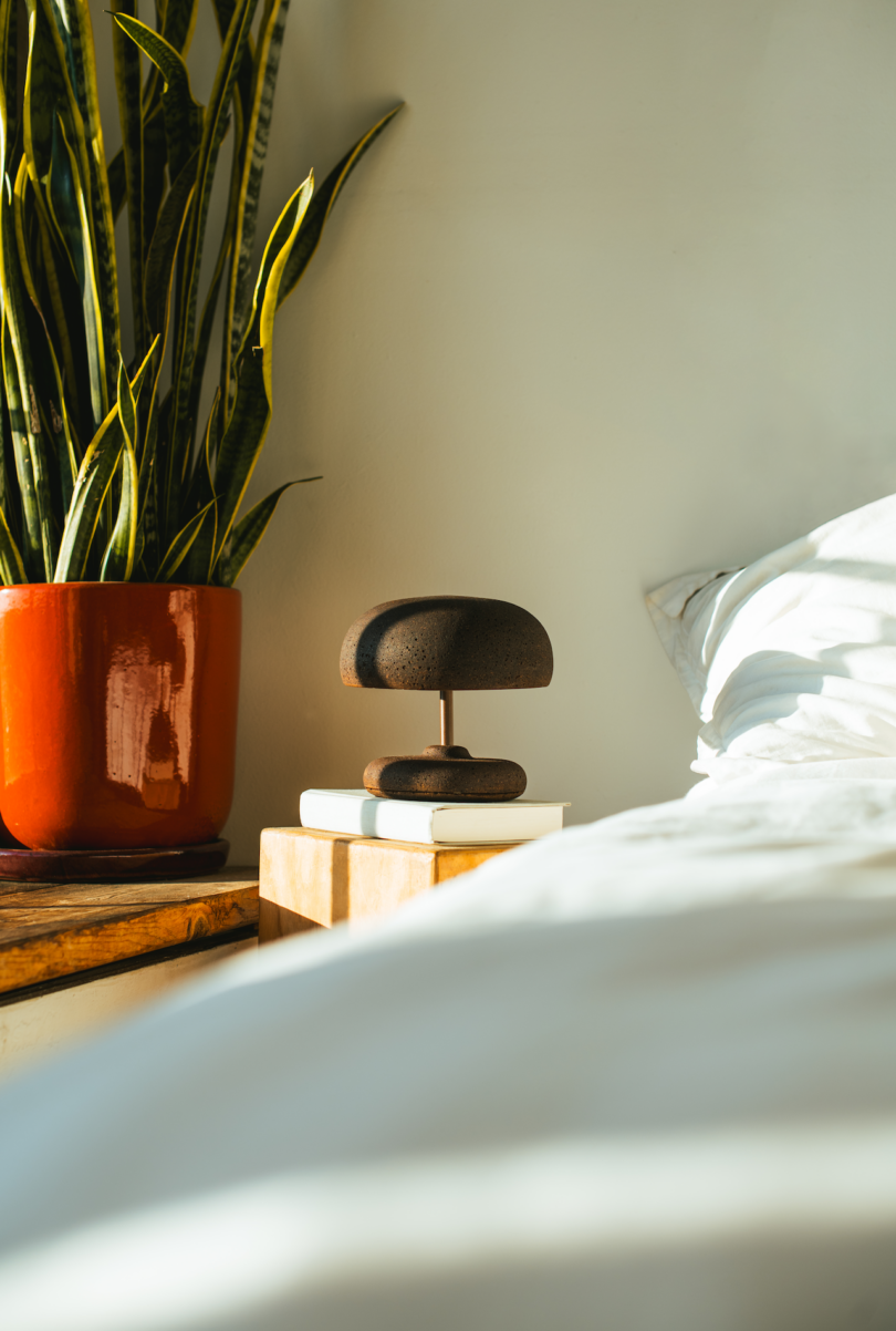 A bedroom scene with a wooden nightstand holding a potted plant, books, and a small lamp. A portion of an unmade bed with white sheets is visible on the right