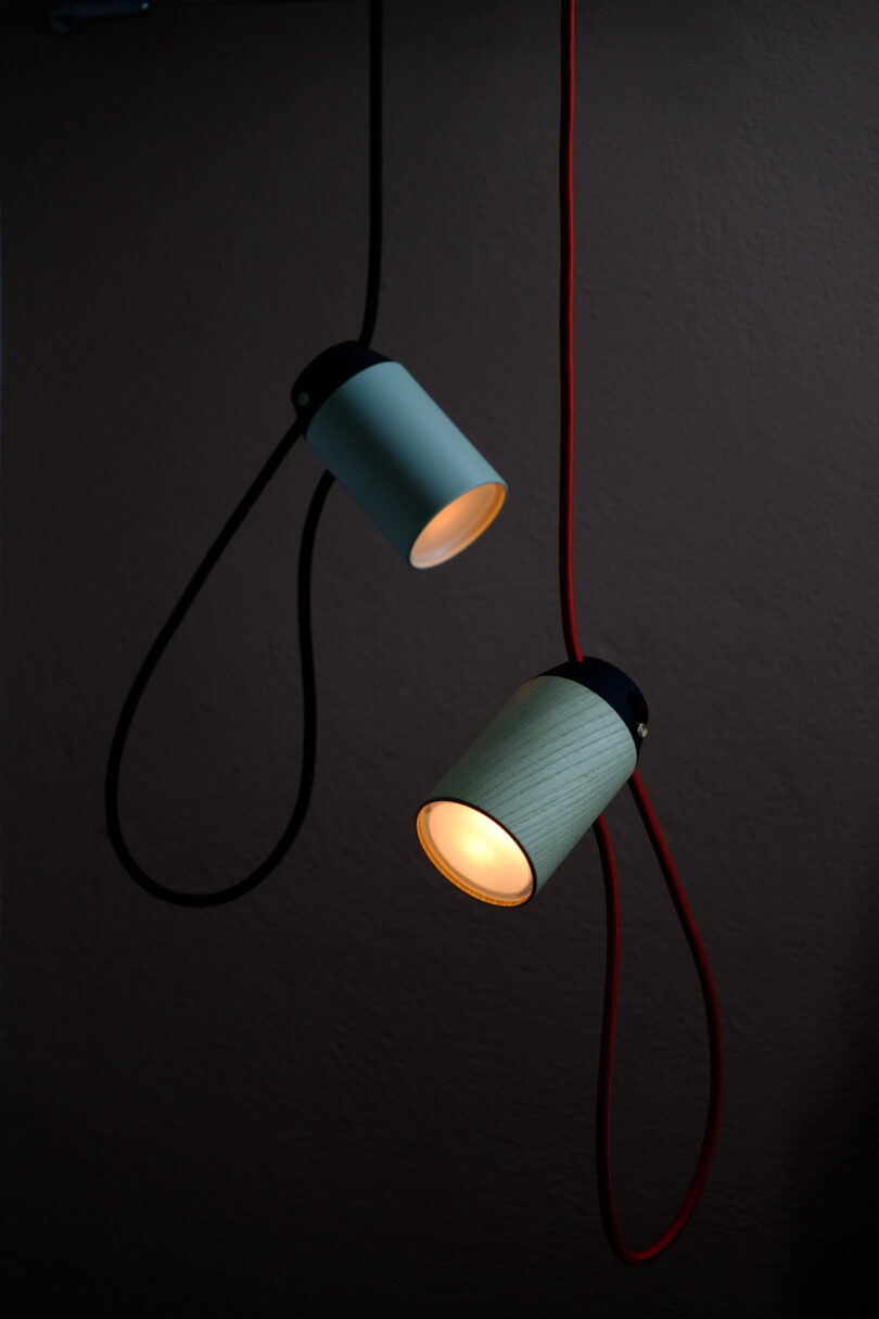 Two cylindrical pendant lights hang against a dark background, one with a black cord and one with a red cord, both emitting a warm glow