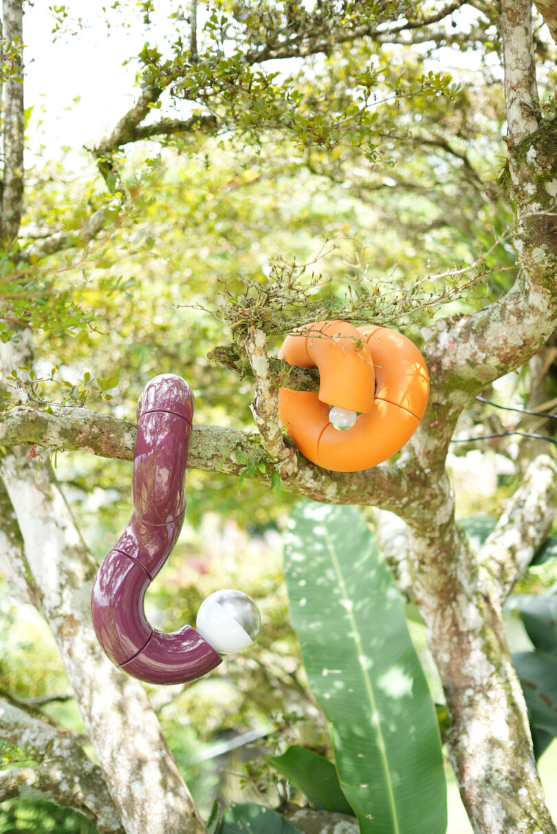 Two abstract sculptures, one purple and one orange, hang from tree branches in a leafy outdoor setting