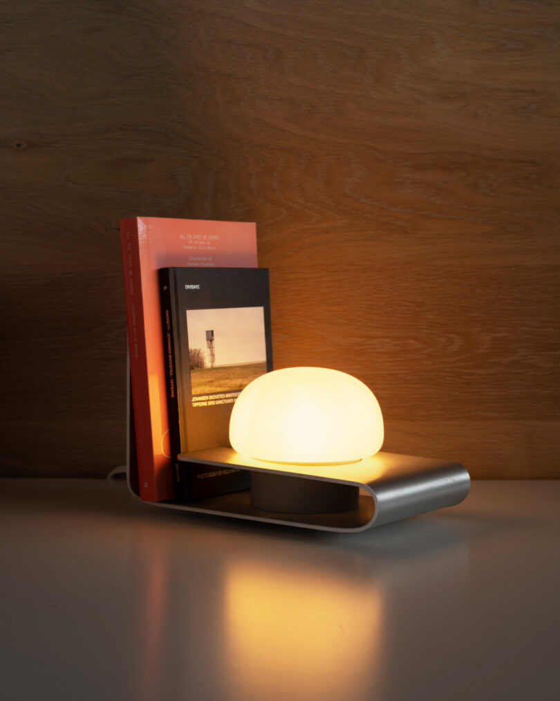 A small, modern table lamp with a glowing orb-shaped light sits next to a few upright books on a gray surface against a wooden background