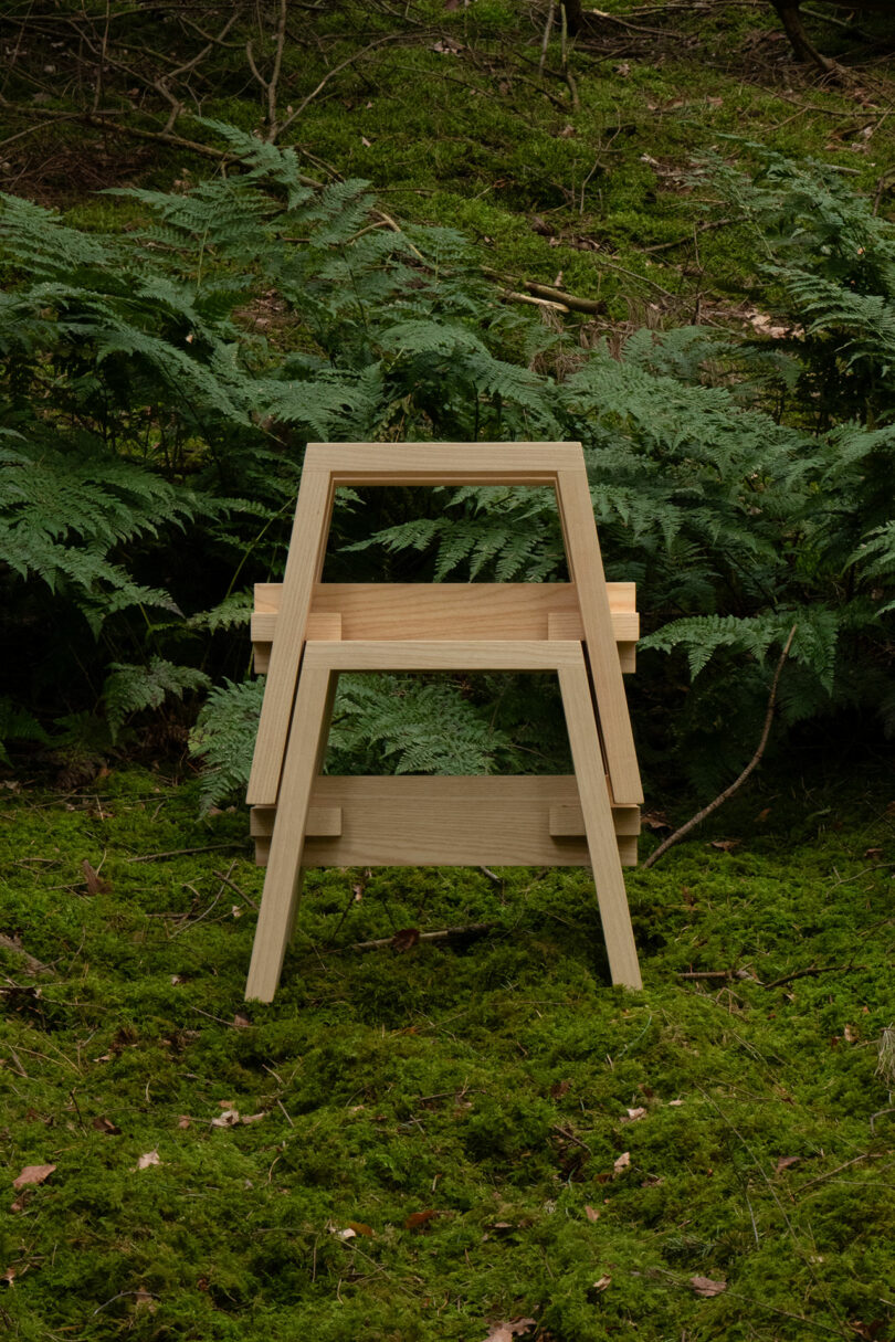 A wooden chair with a minimalist design is placed on a moss-covered forest floor, surrounded by ferns.