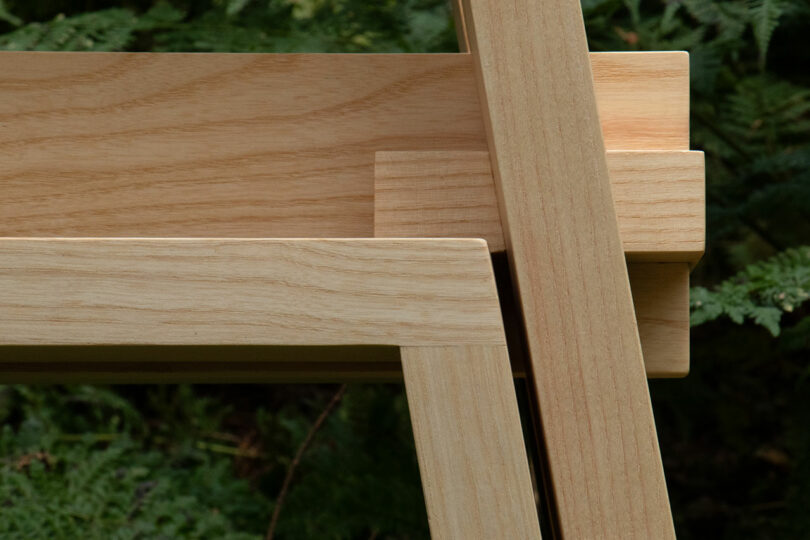 Close-up of a wooden chair with intersecting beams set against a backdrop of green foliage.