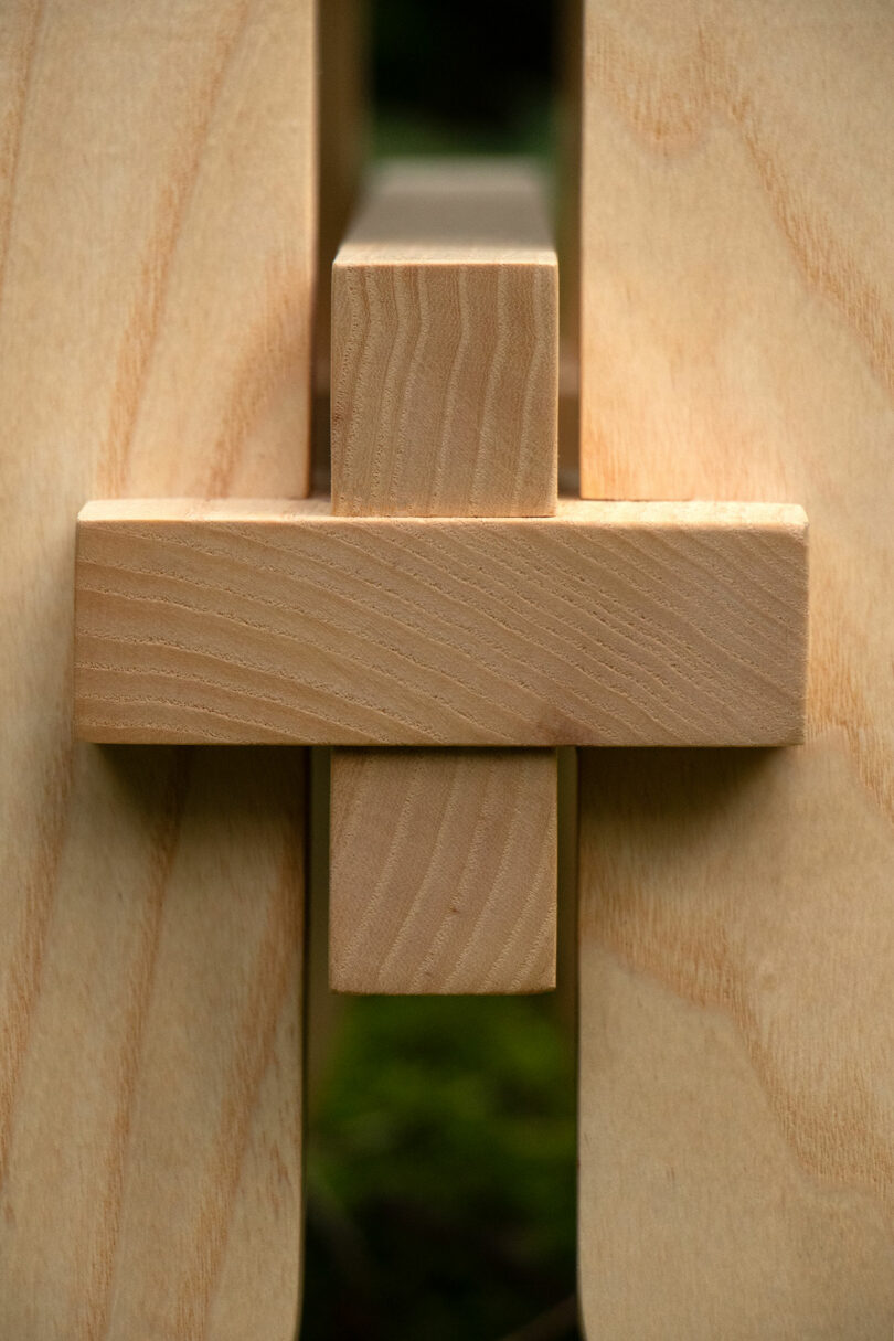 Close-up of interlocking wooden pieces with a light wood grain texture, forming a cross shape against a blurred green background.