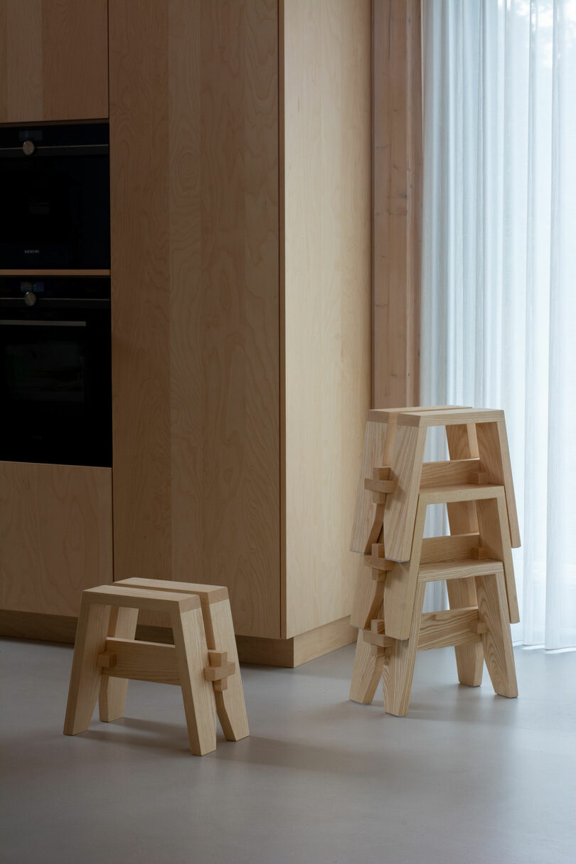 Four wooden stools, one standing alone and three stacked, sit near light-colored wooden cabinets and a sheer curtain in a minimalist room.