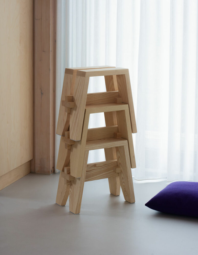 Three wooden stools are stacked in front of a sheer white curtain, with a purple cushion on the floor nearby.