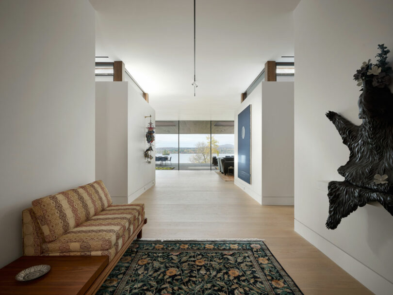 A hallway adorned with a patterned rug, a tapestry sofa, and striking wall art leads to a living room. Large windows at the end offer a breathtaking view of the landscape.