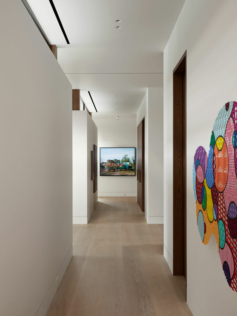 A modern hallway with wooden flooring and white walls showcases vibrant abstract artwork on the right, complemented by a serene landscape picture framed at the end.