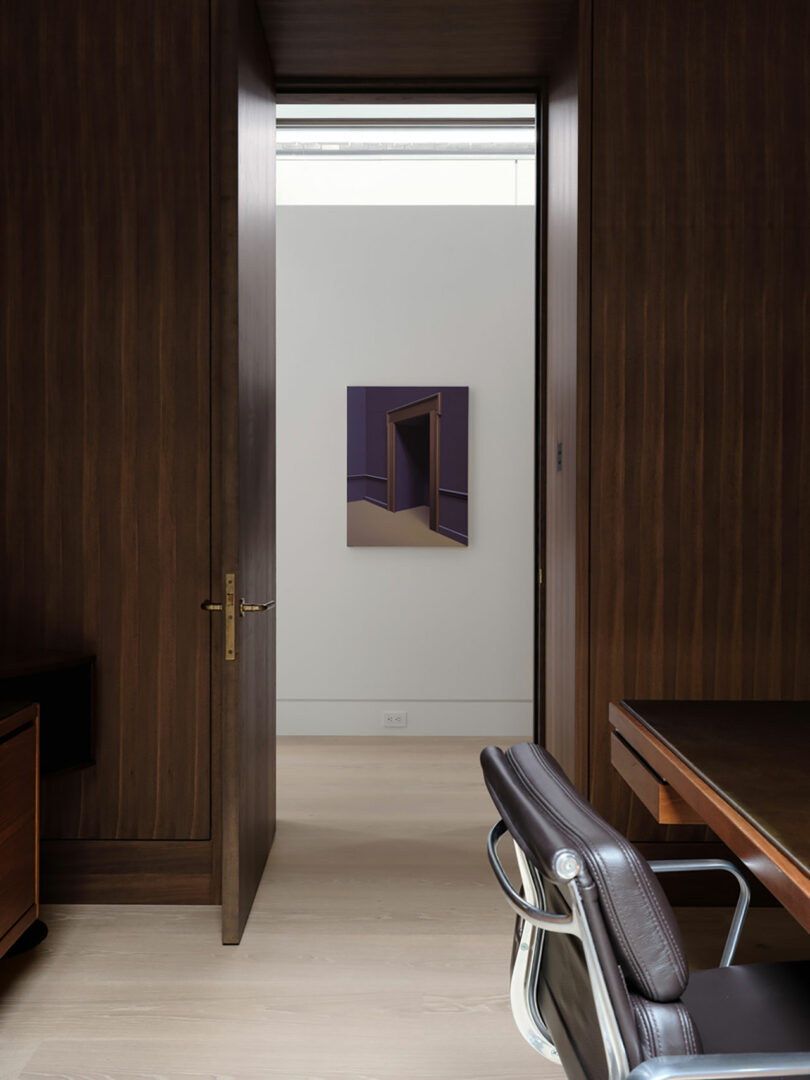 An office with a leather chair and wooden desk. An open door reveals a minimalist painting on a light gray wall.