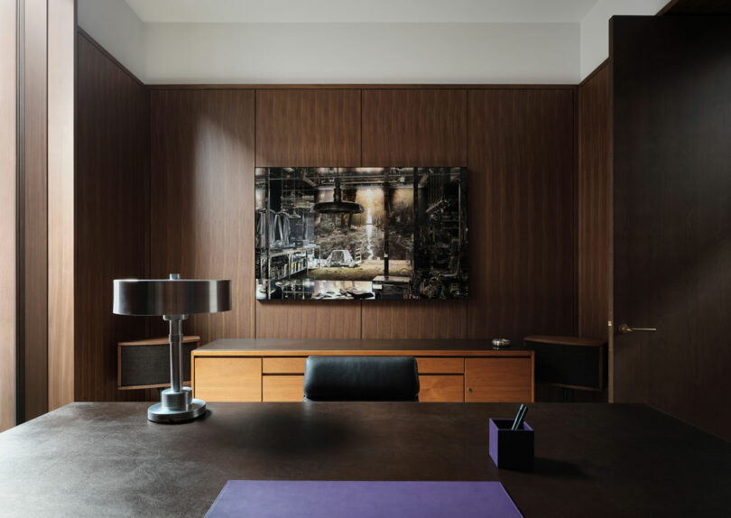 A modern office from Atelier Echelle features a wooden desk, a black leather chair, a metallic lamp, and wall-mounted industrial-themed artwork.