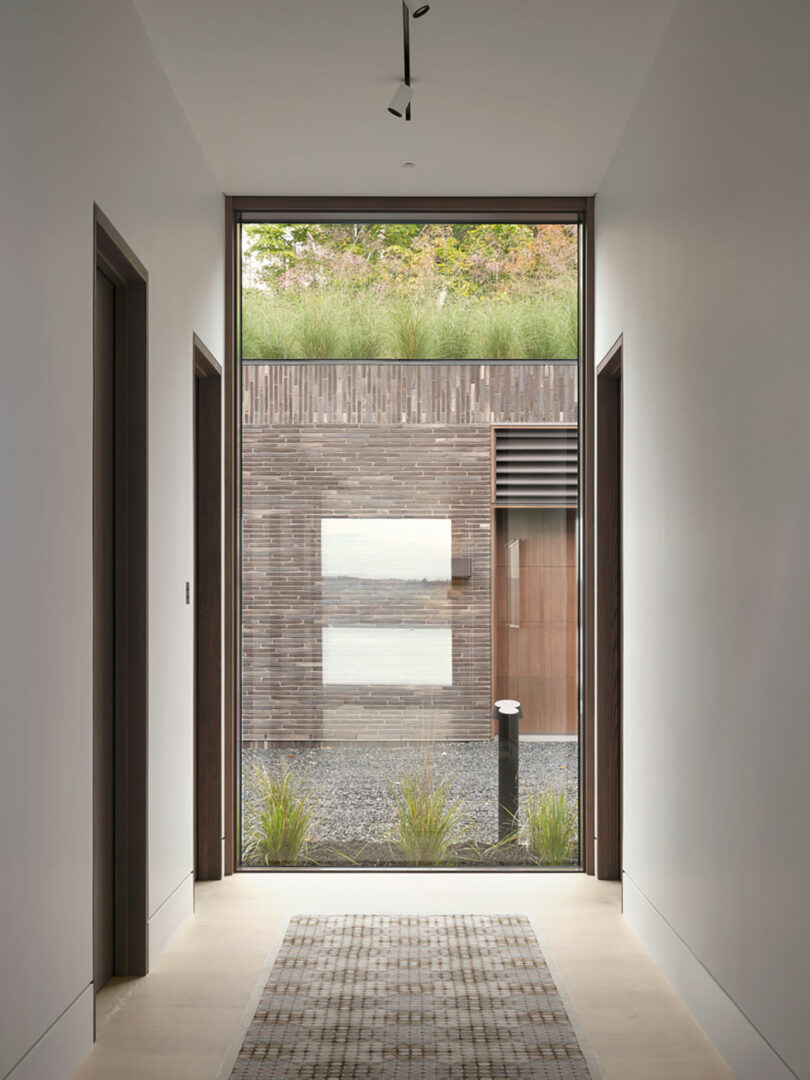 The hallway with its large window offers a view of a modern building designed by Atelier Echelle, showcasing wooden panels and lush greenery on the roof. A doormat lies quietly in the corridor, framing this architectural harmony.