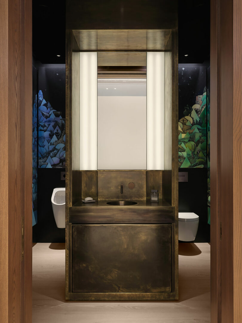 The modern bathroom features a brass sink and large mirror framed by two lit panels, capturing the elegant essence of Atelier Echelle. A colorful abstract mural adds a vibrant touch to the walls, creating a harmonious blend of art and design.