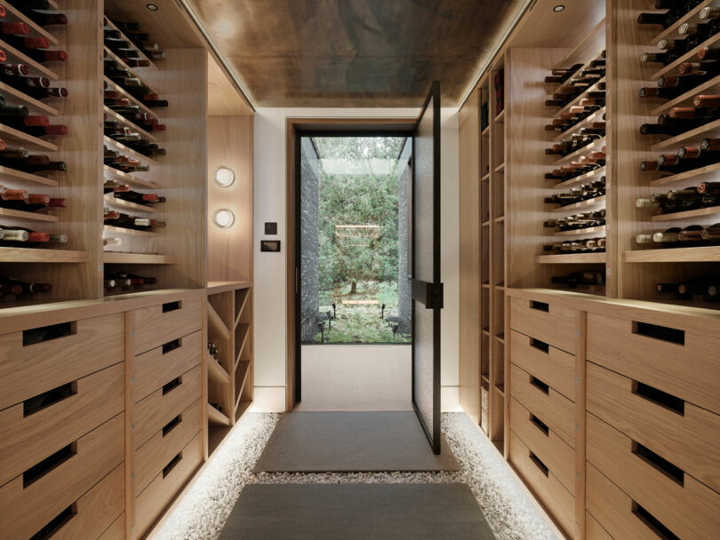 A modern wine cellar designed by Atelier Echelle, with wooden shelves and a glass door, showcases rows of wine bottles and opens to a serene garden view outside.