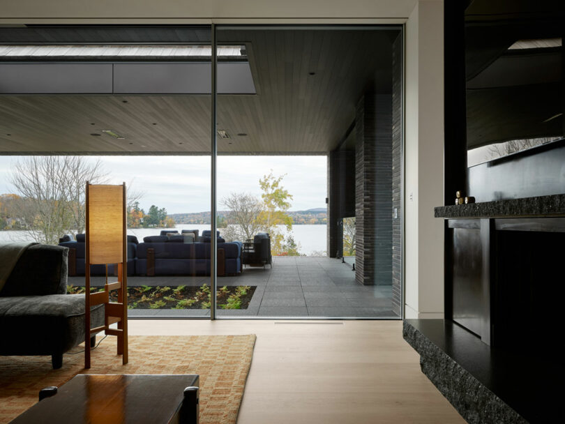 Interior view of a modern living room, designed by Atelier Echelle, with glass doors opening to a patio. The patio has seating and overlooks a serene lake surrounded by trees under a clear sky.