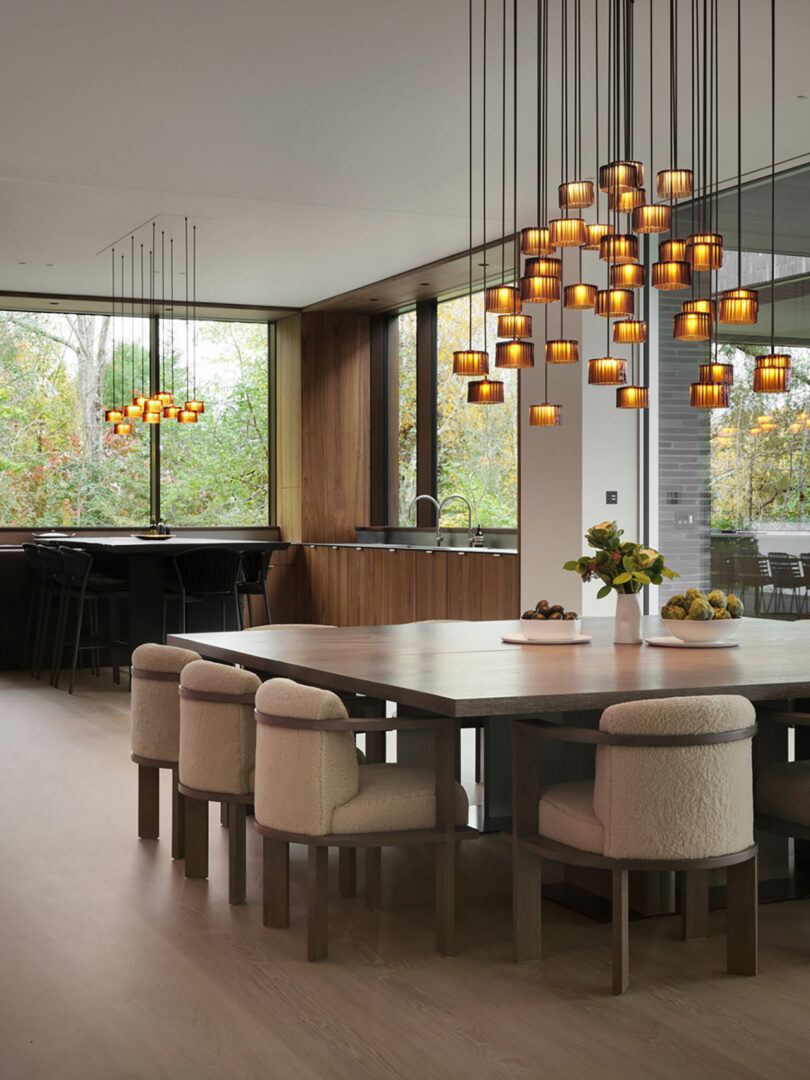 Modern dining area with a large wooden table, cushioned chairs, and pendant lights. The kitchen boasts bar stools in the background, while large windows showcase the trees outside.