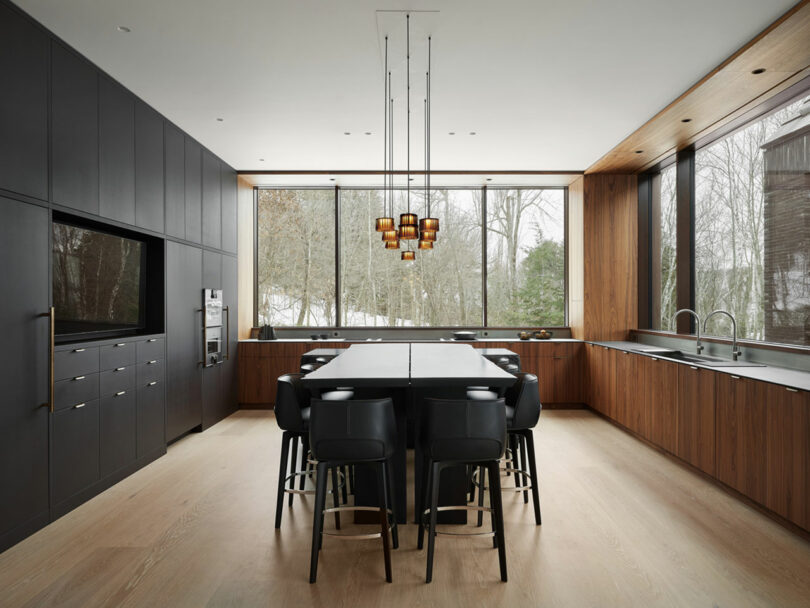 Modern kitchen by Atelier Echelle with dark cabinets, a large island paired with black chairs, pendant lighting, and wide windows showcasing a forest view. Light wood flooring and sleek design elements complete the sophisticated space.