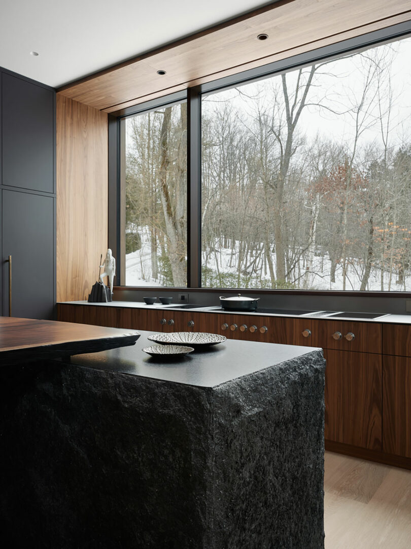 The modern kitchen by Atelier Echelle boasts a large window with views of snowy trees. It features a dark stone island, wooden cabinetry, and minimalist decor, perfectly blending style and nature.