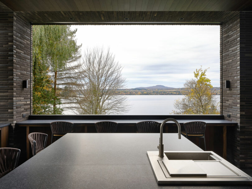 This modern kitchen features a large island and sink that overlook a scenic lake view, surrounded by trees and distant hills.