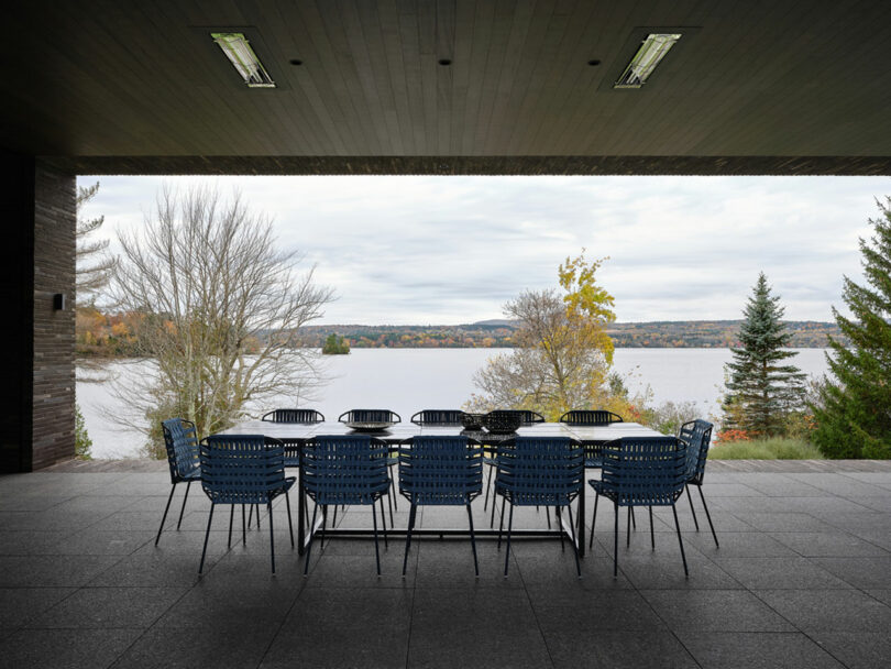 A long table with blue chairs, perfectly set on a covered patio that offers stunning views of a lake embraced by autumn trees.