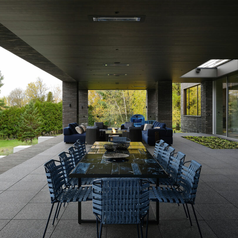 The modern outdoor patio, designed by Atelier Echelle, features a dark ceiling and wicker chairs around a large table. Cozy blue sofas invite relaxation while offering a charming view of the trees in the background.