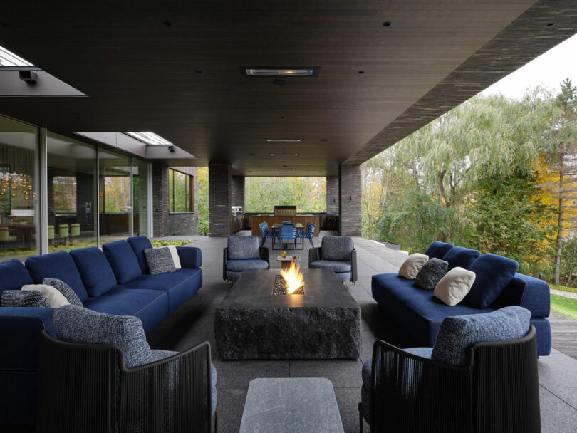 A modern outdoor patio designed by Atelier Echelle features blue sofas around a rectangular fire pit, complemented by a dining area and lush greenery in the background.