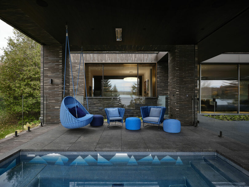 The outdoor patio boasts a shimmering pool and blue chairs, with a hanging chair gently swaying. Glass walls reveal the modern house's chic interior, all enveloped by lush greenery.