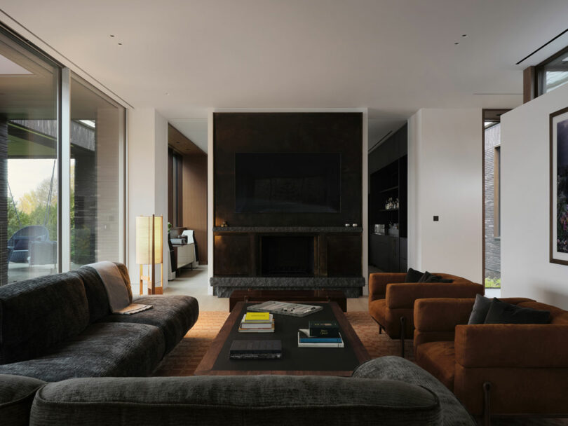 Modern living room with dark sofas, brown armchairs, and a large TV above a fireplace. Floor-to-ceiling windows flood the space with natural light.