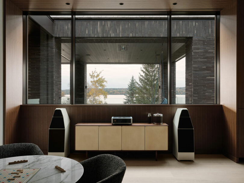 A modern living room designed by Atelier Echelle boasts large windows offering scenic views of trees and water. A sleek console with audio equipment and two speakers complements a round table with chairs in the foreground, creating a harmonious blend of style and nature.