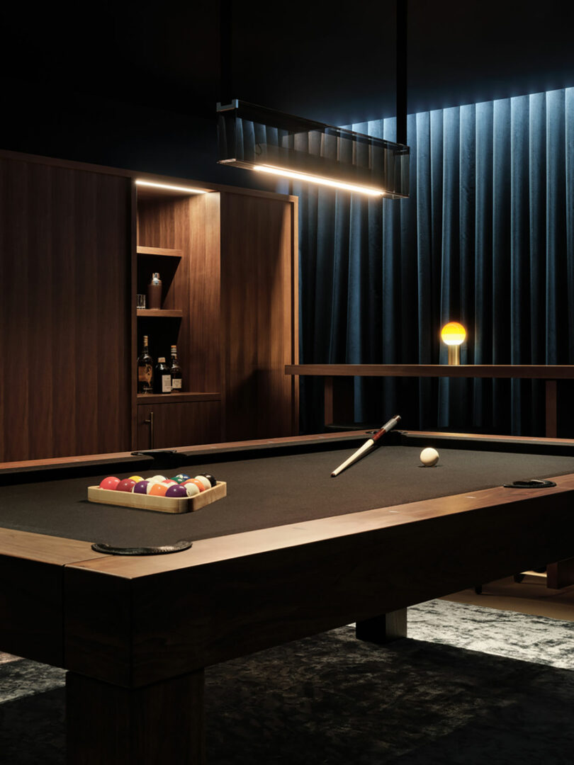 A dimly lit billiard room features a pool table set for a game. Balls and cue are perfectly arranged on the table, while a shelf with bottles and a curtain add to the sophisticated ambiance in the background.