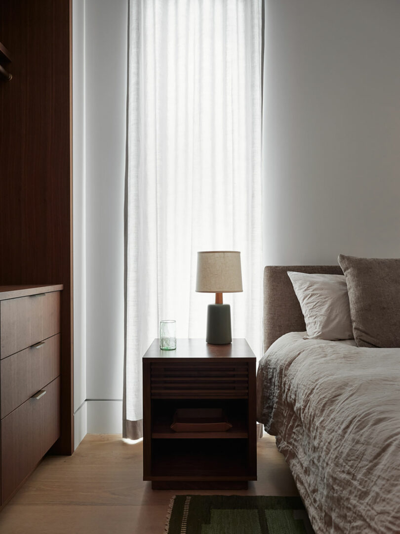 This bedroom features a bed, a wooden nightstand adorned with a stylish lamp and glass, alongside a tall window dressed in sheer curtains.