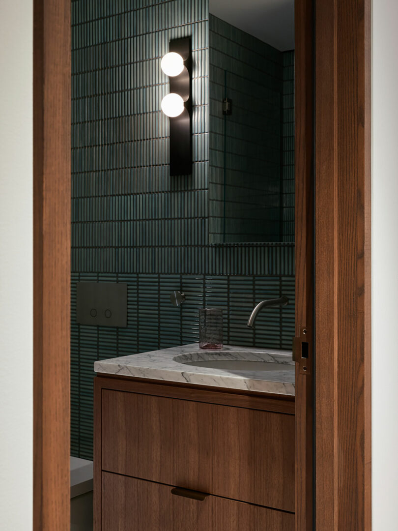 The bathroom features an Atelier Echelle dark wood vanity, complemented by a marble countertop and modern fixtures. Green tiled walls provide a stunning backdrop, highlighted by a vertical black light fixture with two round bulbs above the sink.